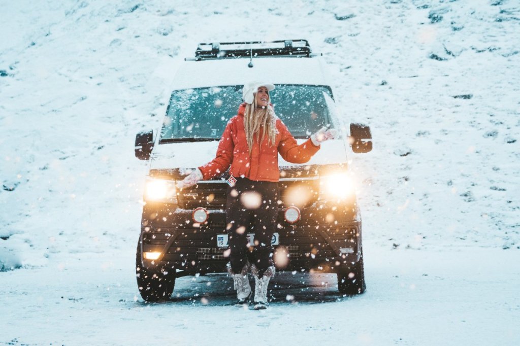 A woman in a red jacket enjoying snowfall in front of a camper van, set against a picturesque Icelandic winter landscape.