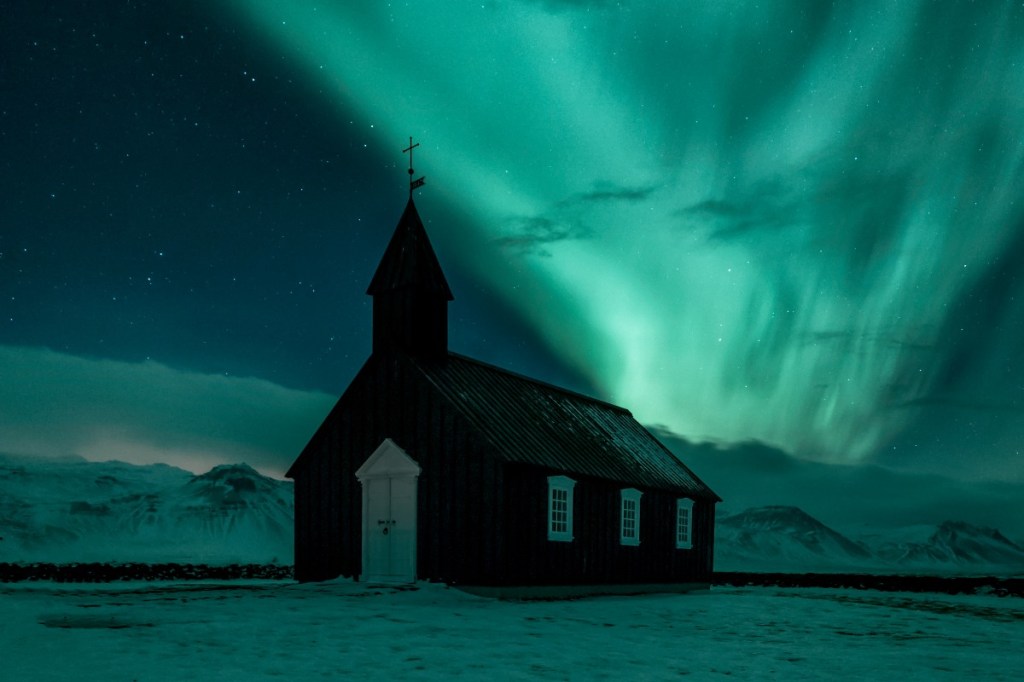 A small black church under the vibrant green Northern Lights in Iceland, surrounded by snowy mountains.