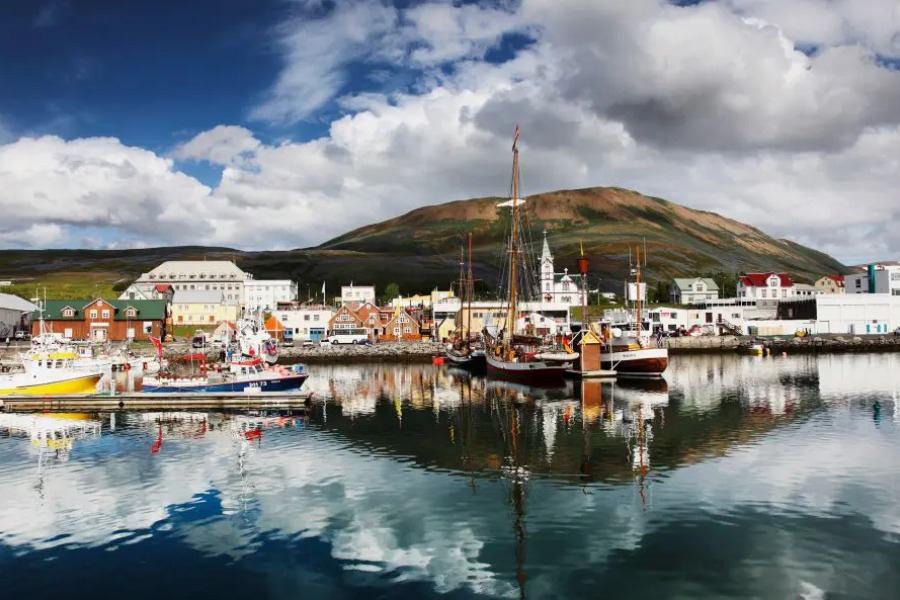 The picturesque harbor of Húsavík in Iceland, with colorful fishing boats, a scenic mountain backdrop, and reflections on the water.