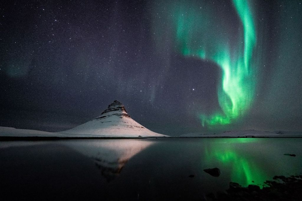 The iconic Kirkjufell mountain in Iceland illuminated by the green glow of the Northern Lights, reflected in a calm lake.