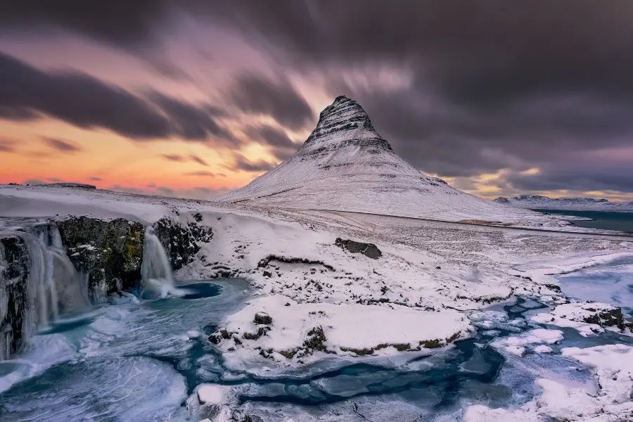 The iconic Kirkjufell mountain during winter, surrounded by frozen waterfalls and dramatic skies with hints of sunset colors.