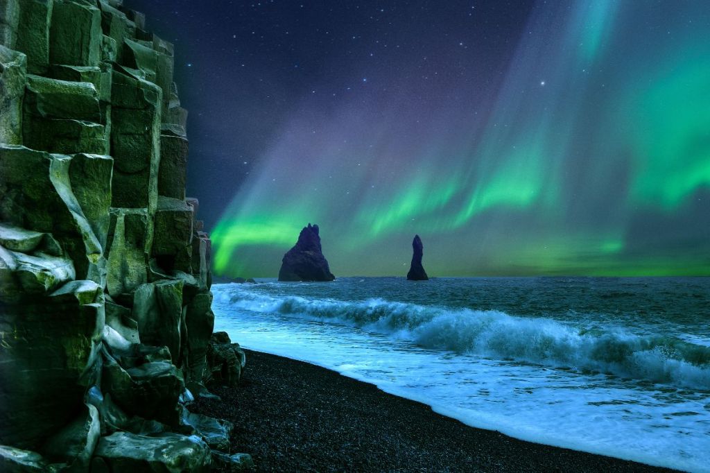 The famous basalt columns and sea stacks of Reynisfjara beach under a stunning display of green Northern Lights in Iceland.