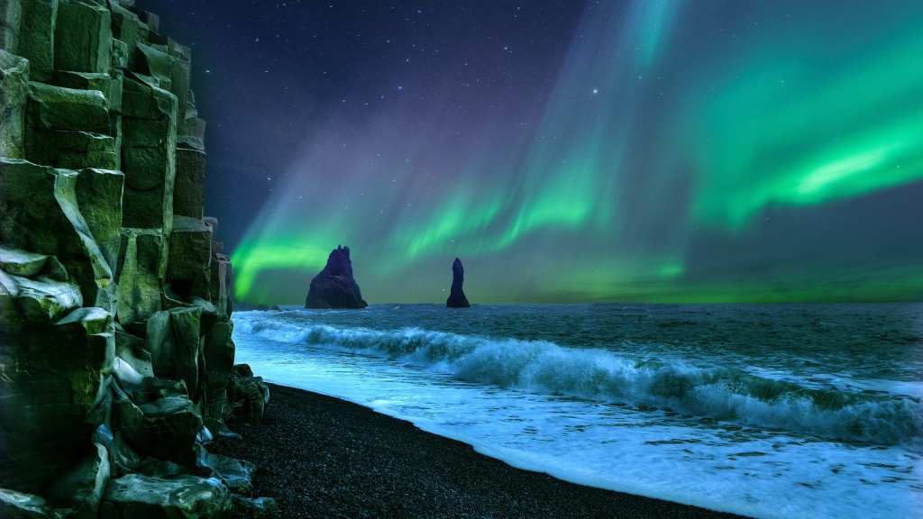 The famous basalt columns and sea stacks of Reynisfjara beach under a stunning display of green Northern Lights in Iceland.