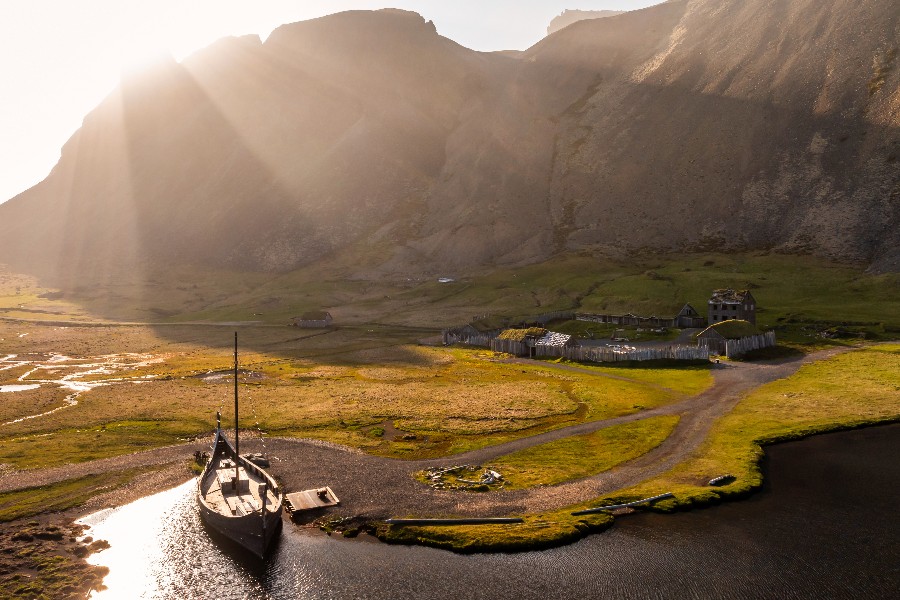 A scenic view of the Viking Village in Stokksnes, Iceland, with a traditional Viking-style ship in the foreground and wooden structures of the replica settlement surrounded by dramatic mountain landscapes illuminated by soft sunlight.