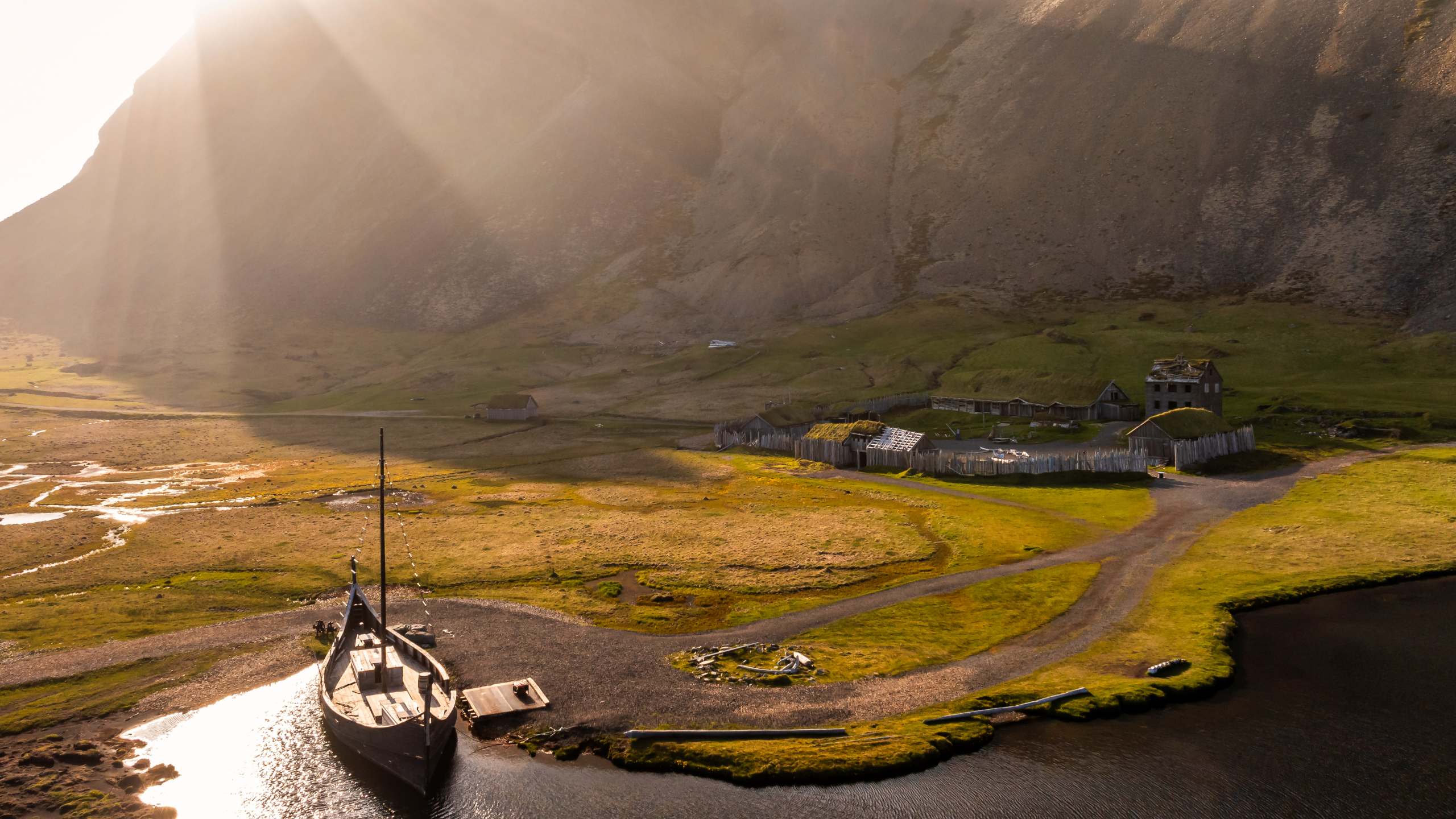 A scenic view of the Viking Village in Stokksnes, Iceland, with a traditional Viking-style ship in the foreground and wooden structures of the replica settlement surrounded by dramatic mountain landscapes illuminated by soft sunlight.