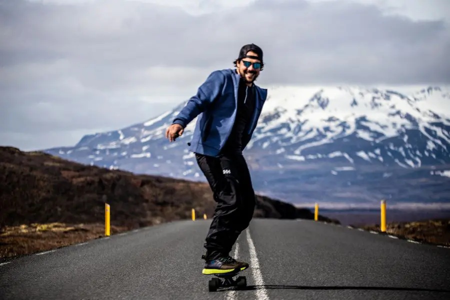 A man enjoying a ride on a longboard on an open road in Iceland, with snowy mountains in the background under a partly cloudy sky.