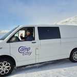 A man wearing sunglasses and a beanie waves from the driver’s seat of a white Camp Easy van parked on a snowy road with mountains in the background.