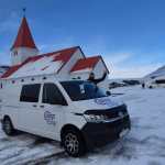 A white Camp Easy camper van parked in front of a snow-covered church with a red roof, as a person waves from behind.