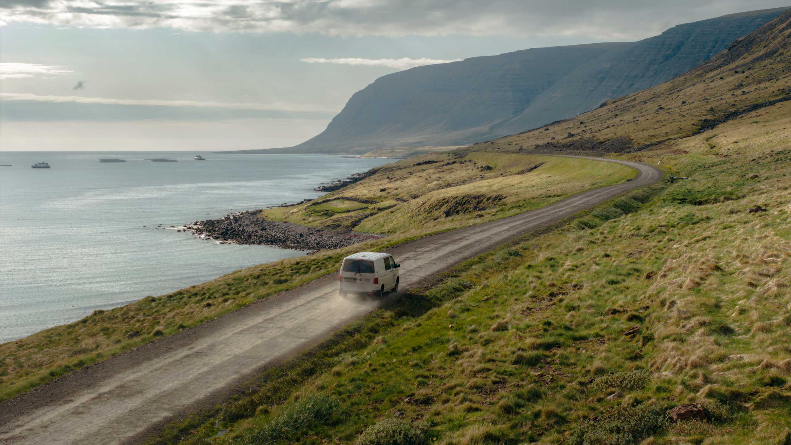 Campervan driving on rocky road next to the shore and green fields
