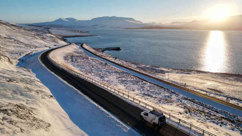 White campervan is driving on the road surrounded by winter landscape near the fjord