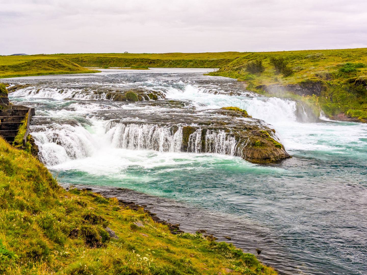 A tranquil waterfall cascades over moss-covered rocks into a vibrant blue river, surrounded by lush green grass under a cloudy sky. The scene is peaceful and natural.