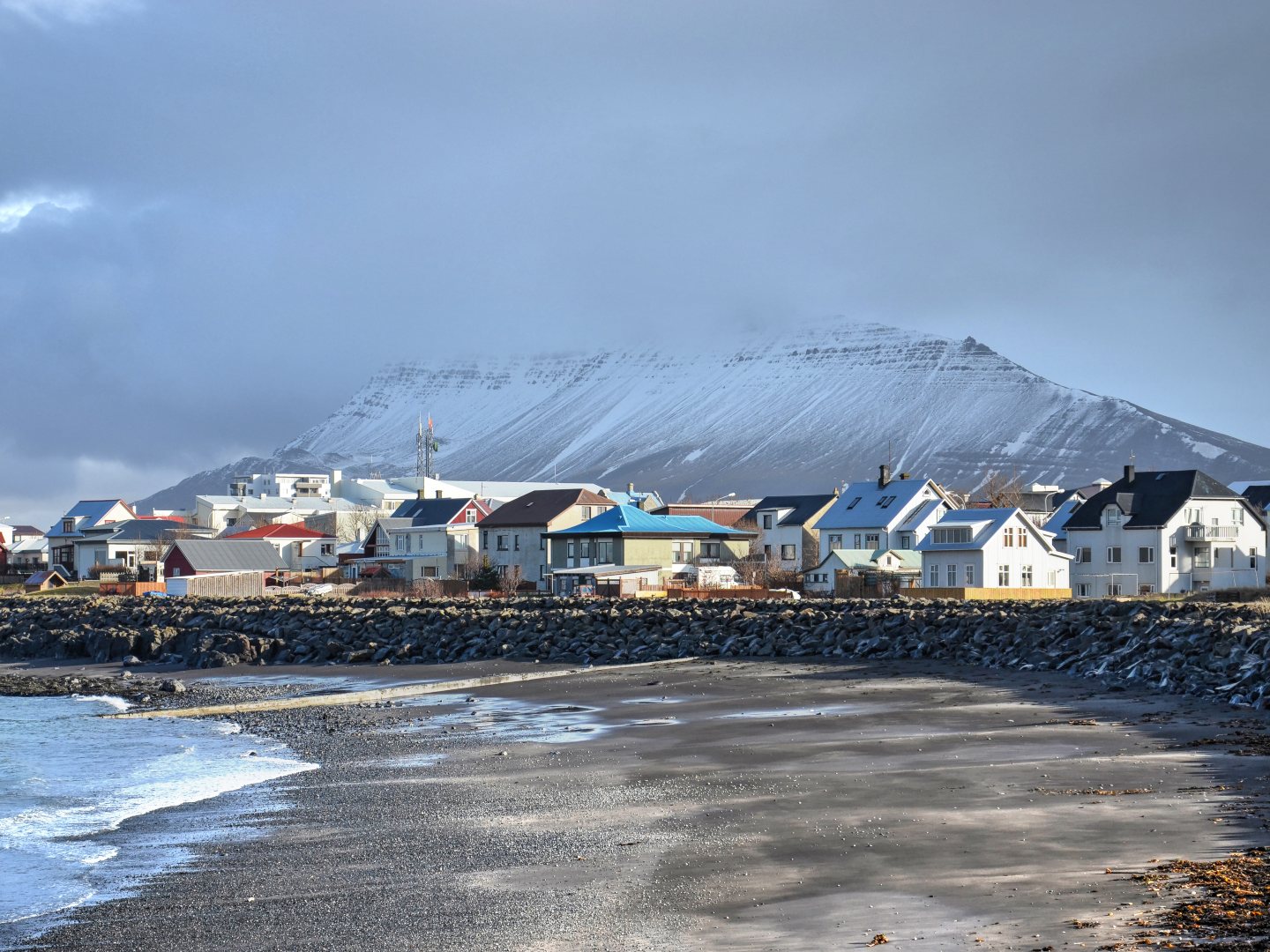 A coastal town with colorful houses sits at the foot of a snow-capped mountain under a cloudy sky, with a calm seashore in the foreground.