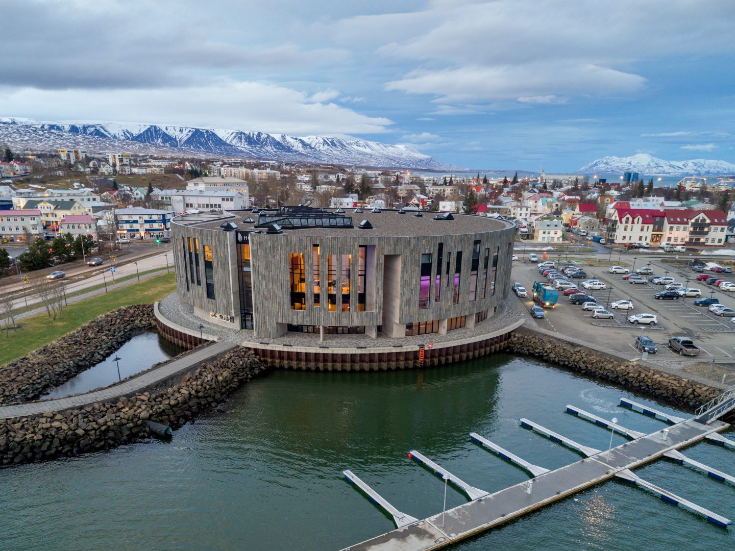 A modern, circular building with large glass windows sits by a waterfront, surrounded by a quaint town and snow-capped mountains under a cloudy sky.