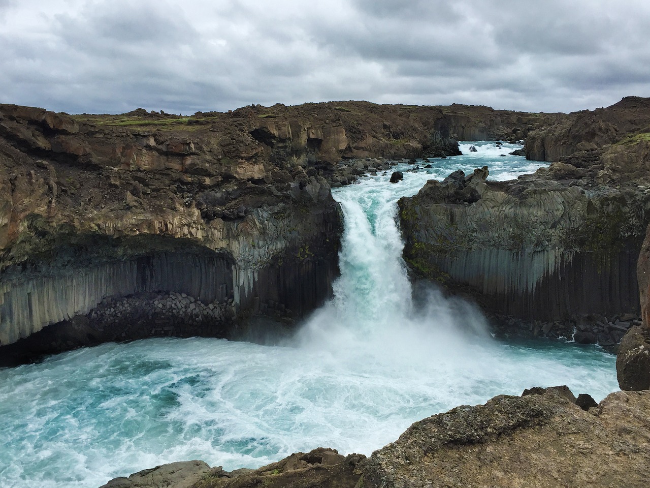 A powerful waterfall cascades over rocky cliffs into a turquoise pool, surrounded by rugged terrain under a cloudy sky, evoking a sense of awe.