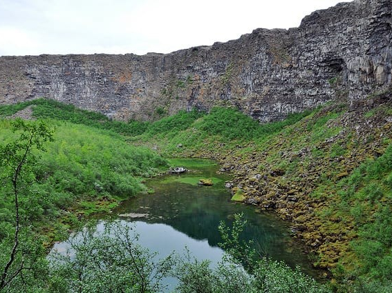 A serene landscape featuring a clear green lake surrounded by lush greenery and rocky cliffs under a cloudy sky. The scene conveys tranquility.