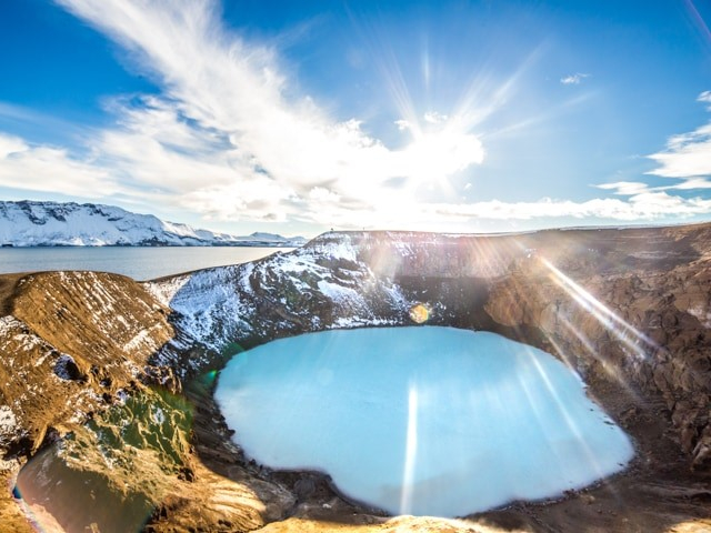 A serene ice-covered crater lake is surrounded by snow-dusted hills under a bright, sunlit blue sky, creating a peaceful and majestic atmosphere.