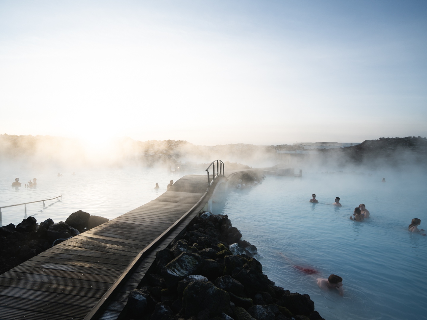 People relax in a steaming outdoor geothermal spa surrounded by rugged landscape at sunrise. A wooden bridge leads across the serene blue water.
