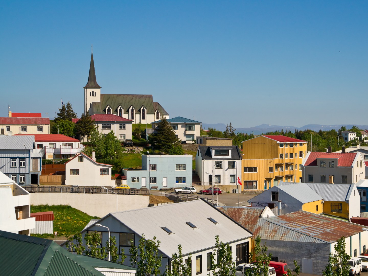 A quaint town with colorful houses, a prominent church steeple, and lush greenery on a sunny day. The scene conveys a peaceful, picturesque atmosphere.