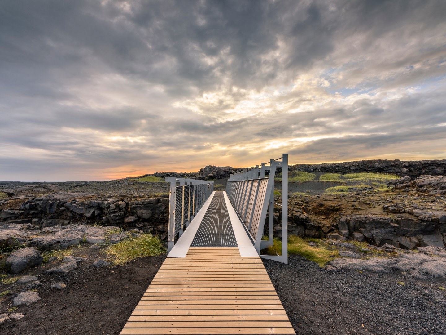 A wooden bridge stretches across rocky terrain under a dramatic, cloudy sky at sunset, evoking a sense of tranquility and exploration.