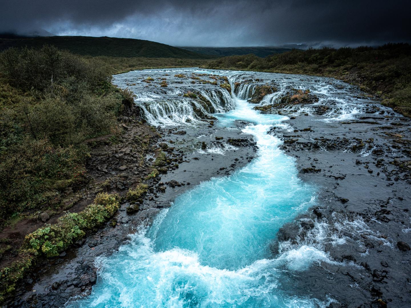 A vibrant blue river cascades over a rocky waterfall amid lush greenery, under dark, stormy clouds, creating a dramatic and serene landscape.