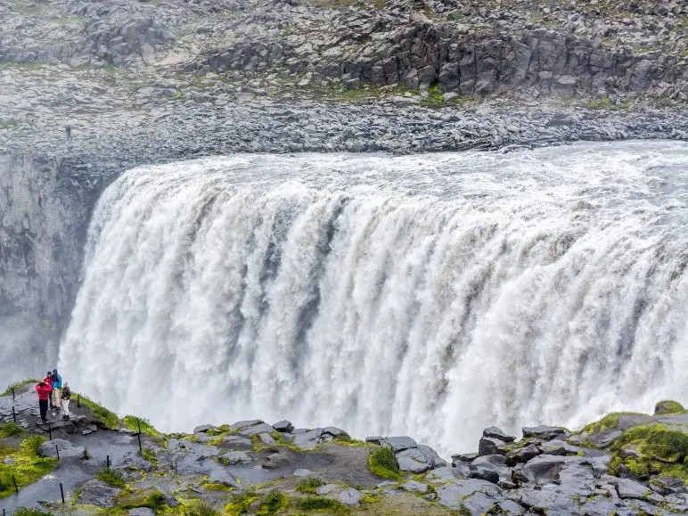 Massive waterfall cascading powerfully over rocky cliffs with mist rising. Three figures in colorful jackets stand on a path, capturing the scene. Dramatic and awe-inspiring.