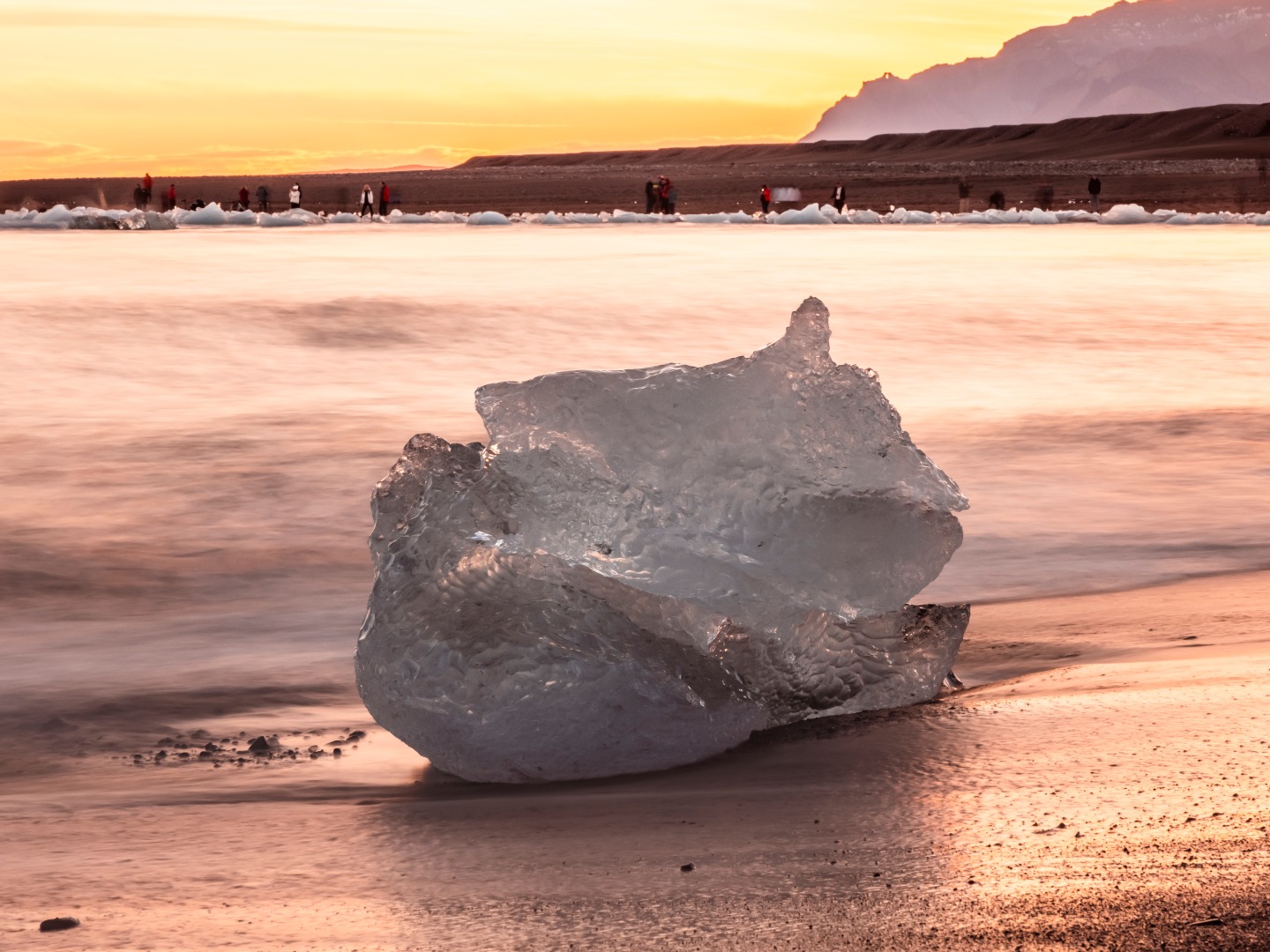 A large ice chunk sits on a black sand beach, reflecting the warm hues of a sunset. Silhouettes of people and distant mountains complete the serene, picturesque scene.