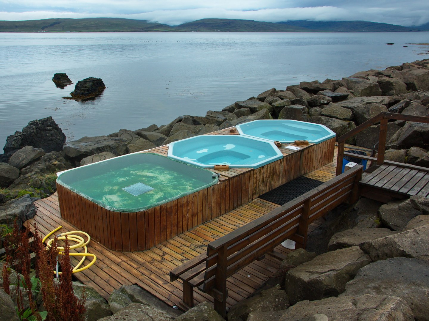 Three wooden hot tubs filled with clear water sit on a rocky shore, overlooking a calm lake. The scene is serene and inviting with a wooden deck.