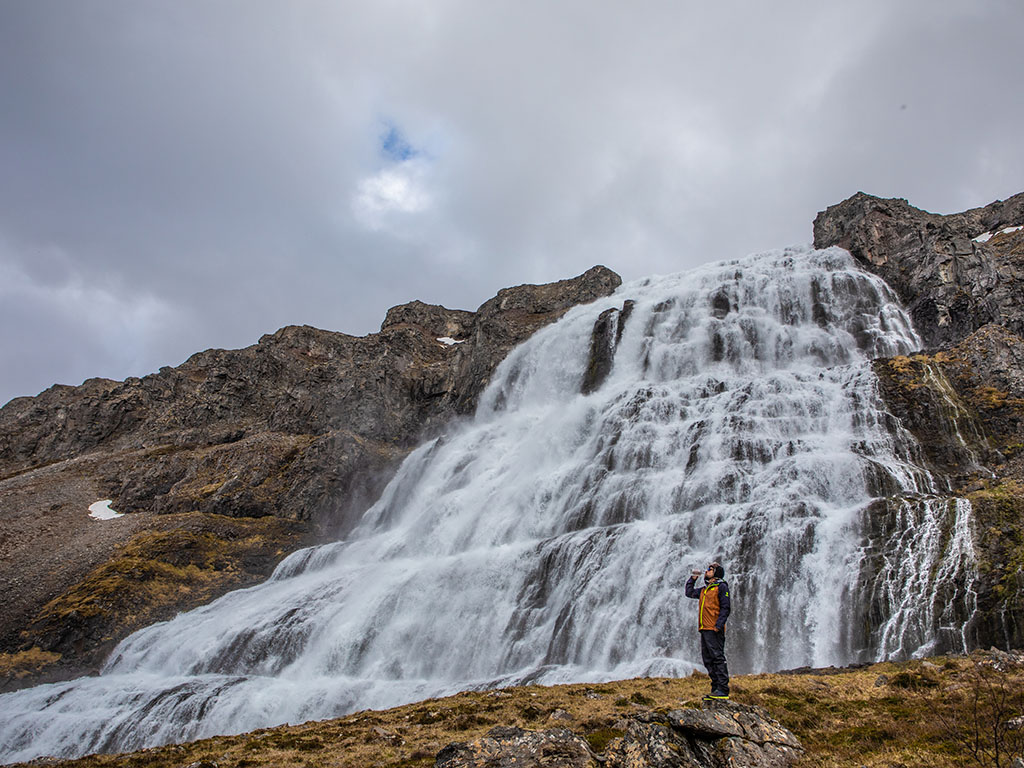 A person in an orange jacket photographs a large, cascading waterfall on a rocky hillside under a cloudy sky, conveying a sense of awe and adventure.