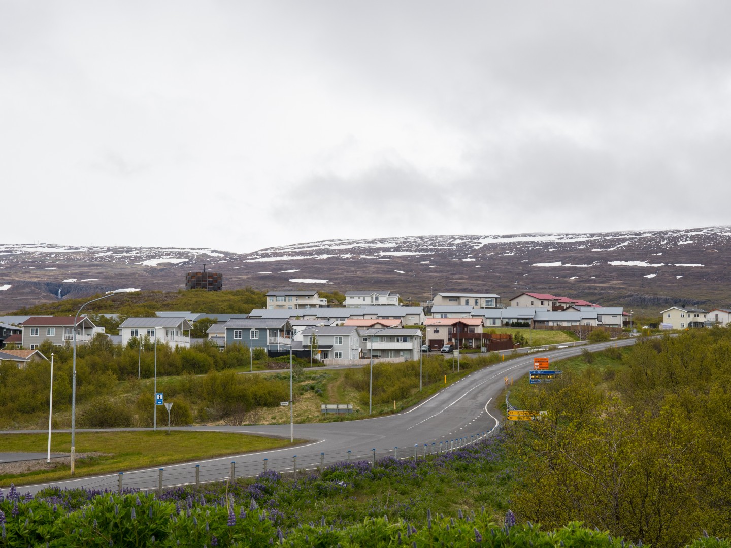 A small village with colorful houses sits under a cloudy sky, bordered by a winding road and green shrubs, with snowy mountains in the background.