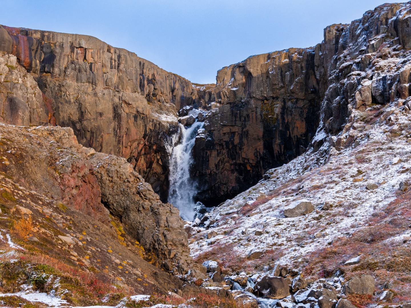 A serene waterfall cascades down rugged, snow-dusted cliffs into a rocky terrain. The scene conveys a tranquil and majestic atmosphere.