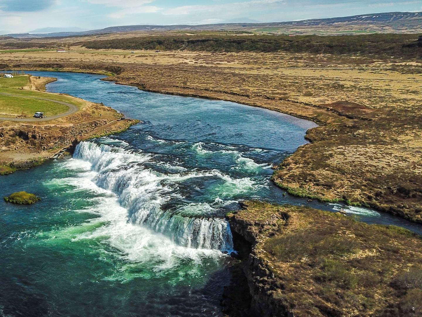 Aerial view of a serene waterfall cascading into a clear blue river, surrounded by grassy fields and a rugged landscape under a partly cloudy sky.