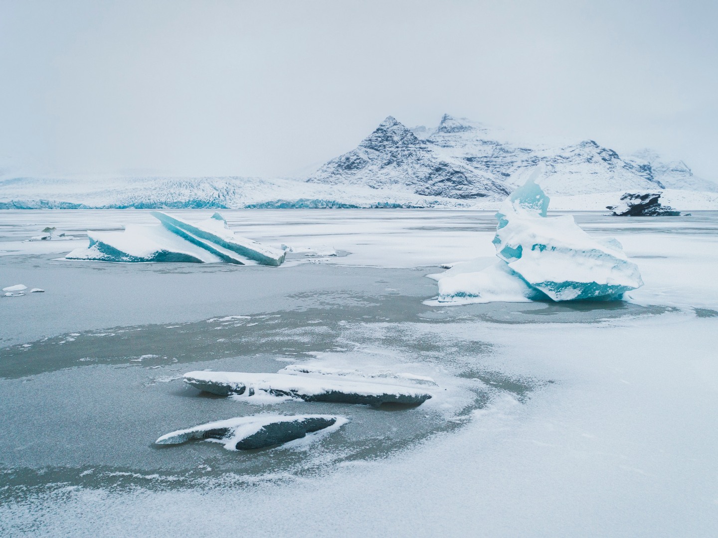 A serene icy landscape with scattered icebergs on a frozen lake, backed by snow-covered mountains under an overcast sky. The scene feels calm and cold.