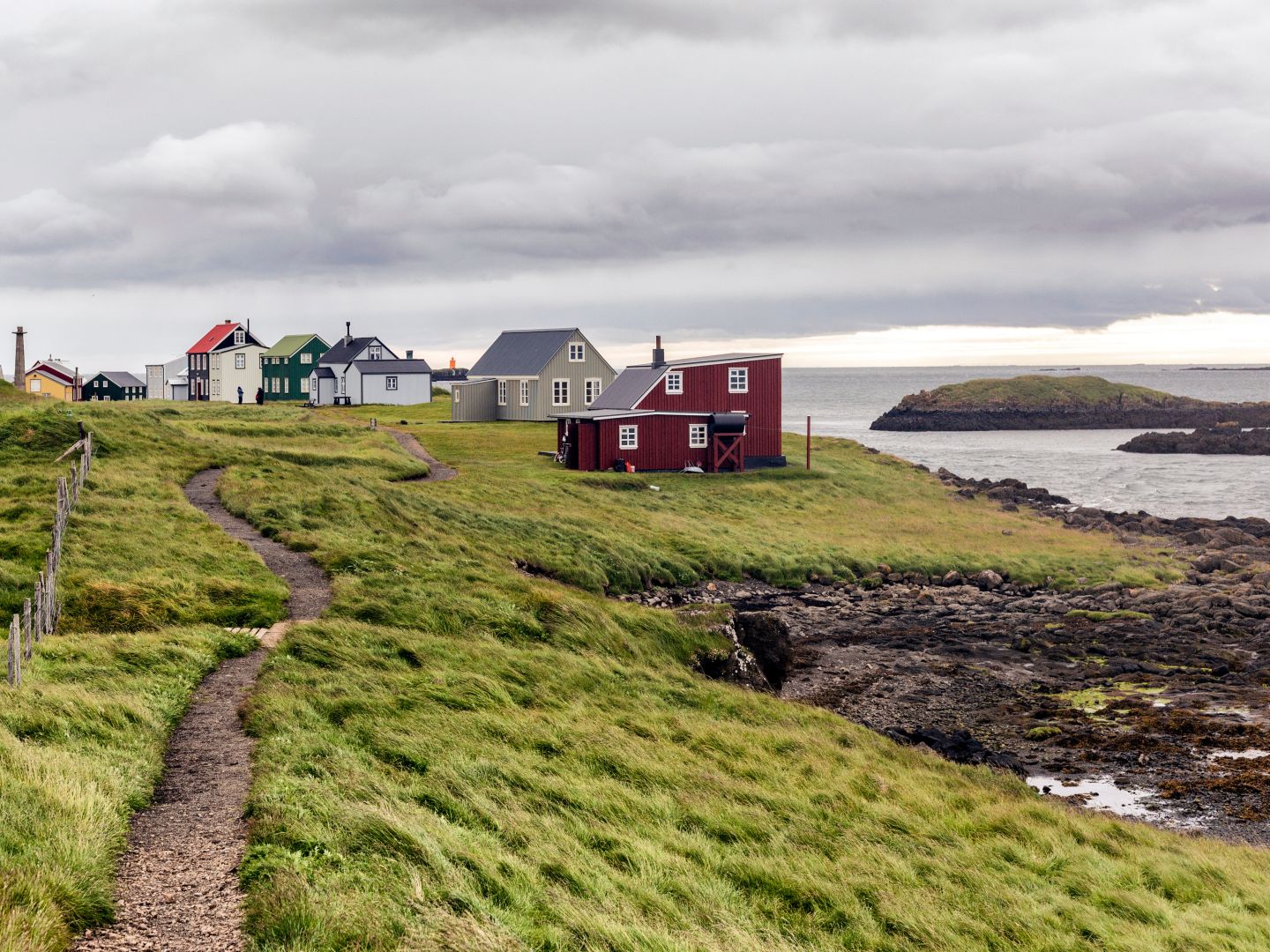 A scenic coastal landscape with colorful houses on a grassy hill under a cloudy sky. A winding path leads through the green terrain, conveying tranquility.
