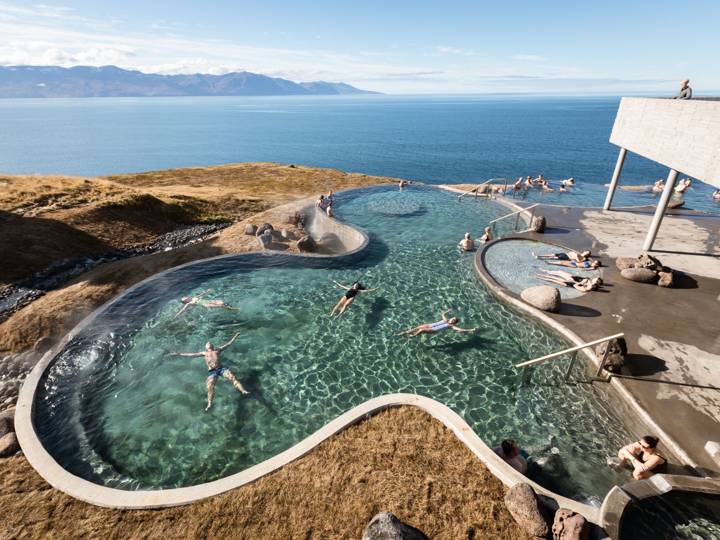 Scenic infinity pool overlooking a vast ocean and distant mountains. People relax in the water under a clear blue sky, conveying tranquility and leisure.