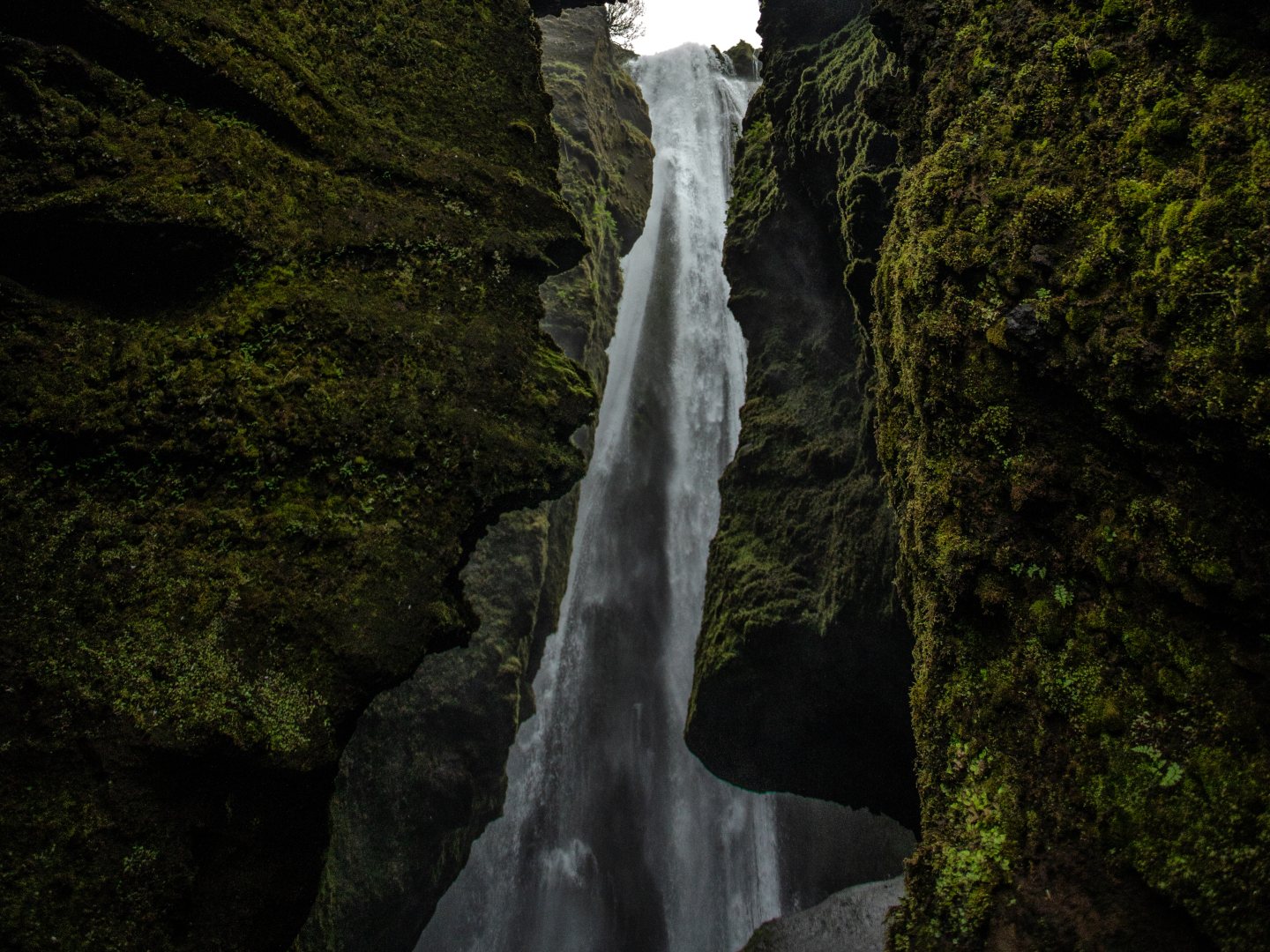 Tall waterfall cascading between moss-covered cliffs, creating a mystical, serene atmosphere. The water flows into a shadowy crevice below.