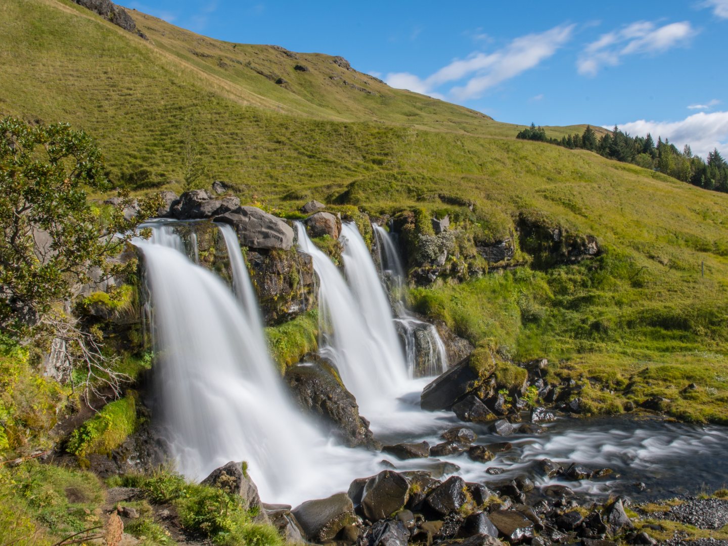 A serene landscape with two cascading waterfalls flowing over dark rocks, surrounded by lush green hills under a bright blue sky with scattered clouds.