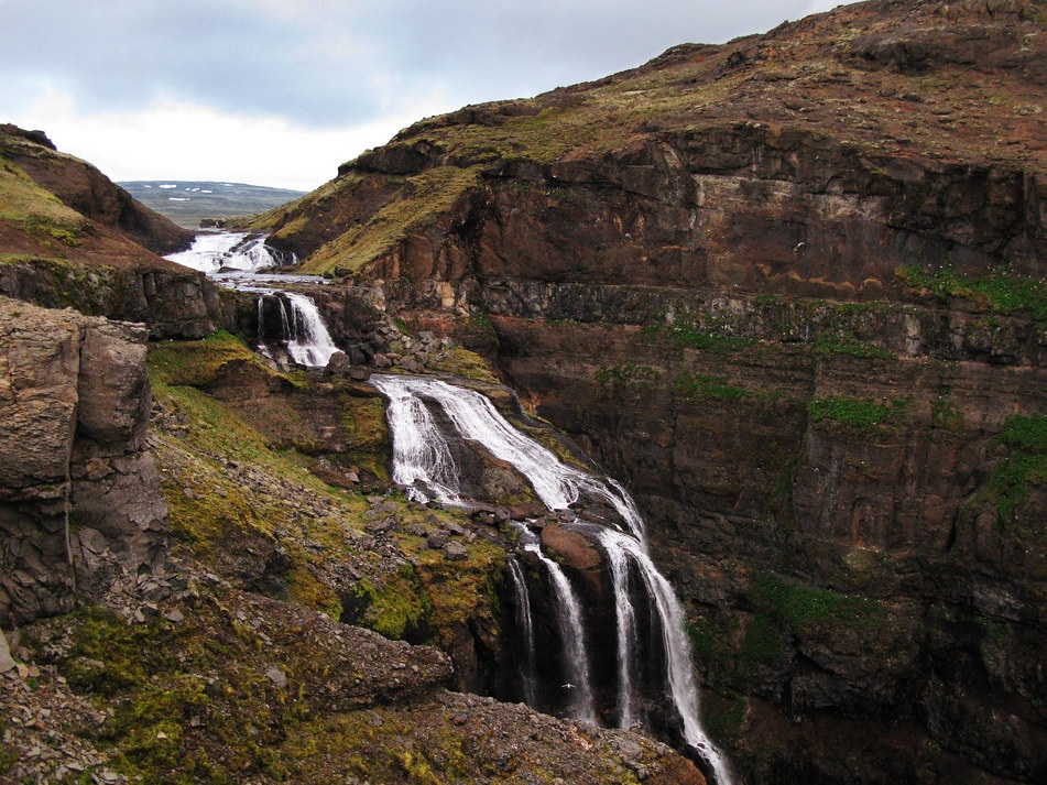 Cascading waterfall flows down a moss-covered rocky cliff in a rugged canyon under a cloudy sky, conveying a serene and majestic natural scene.