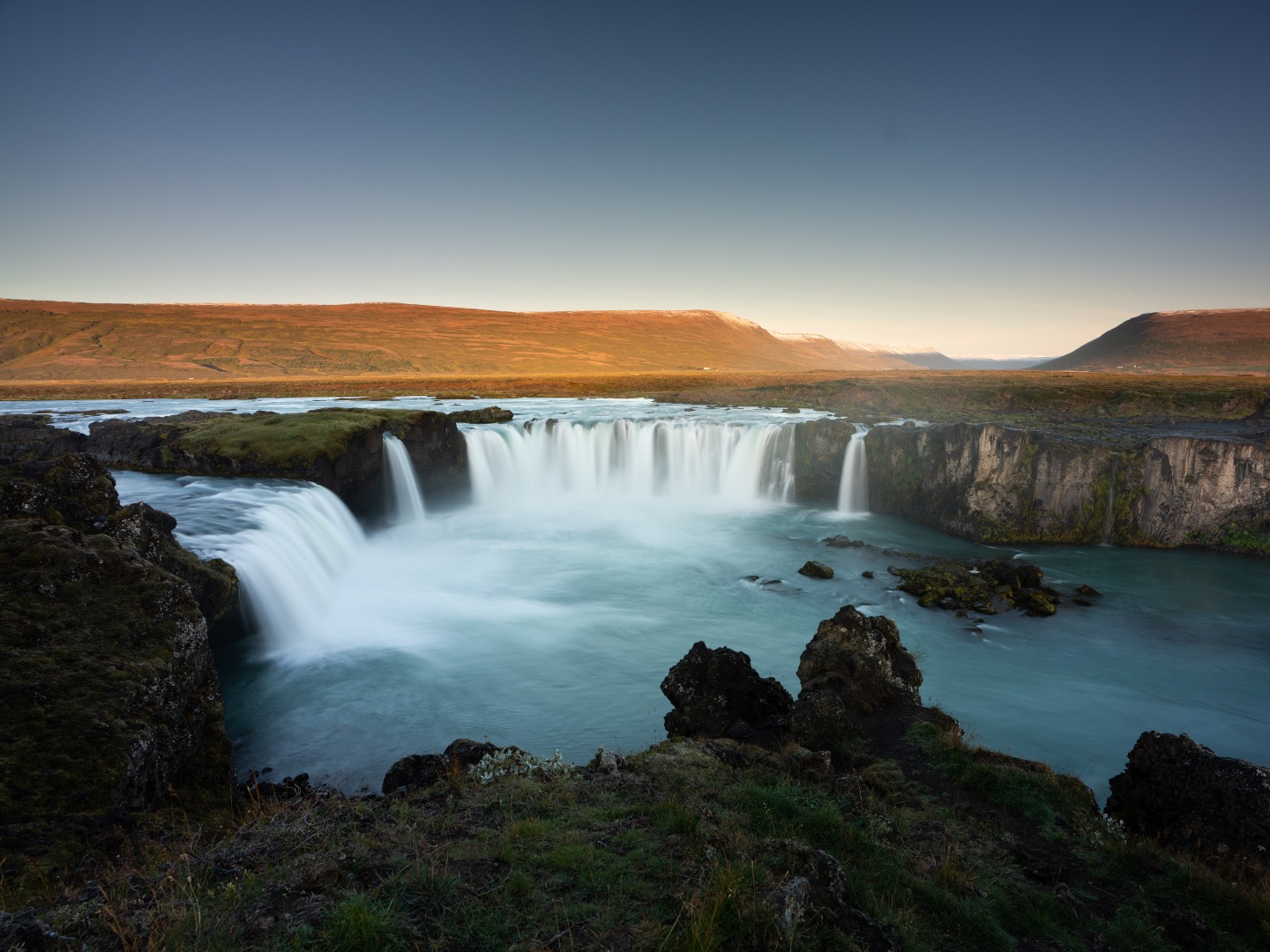 A serene waterfall cascades into a turquoise pool surrounded by moss-covered rocks under a clear sky. The scene evokes tranquility and natural beauty.
