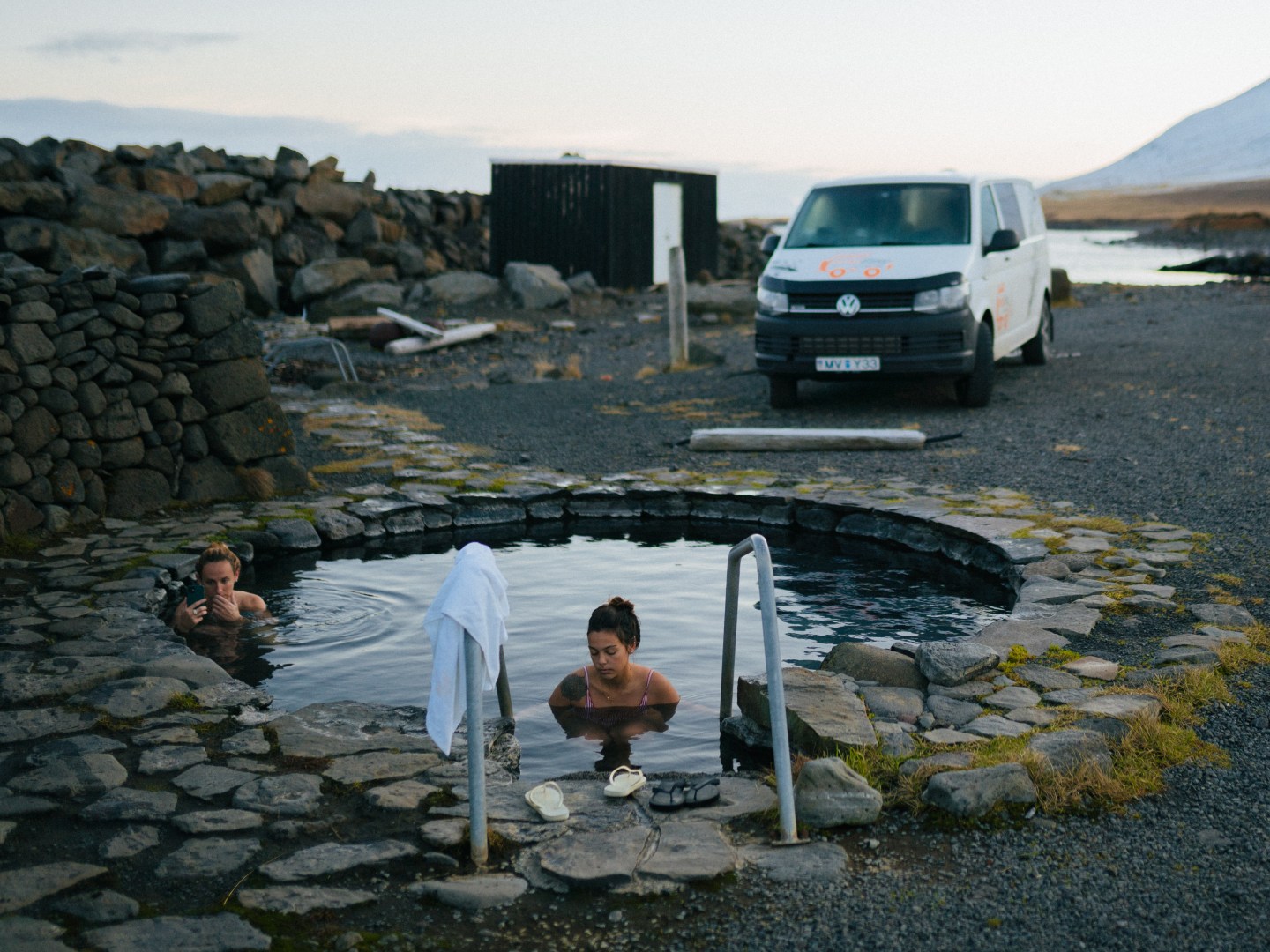 Two people relax in a small, circular hot spring surrounded by stones. A white van is parked nearby on a gravel surface, with snow-capped mountains in the distance.