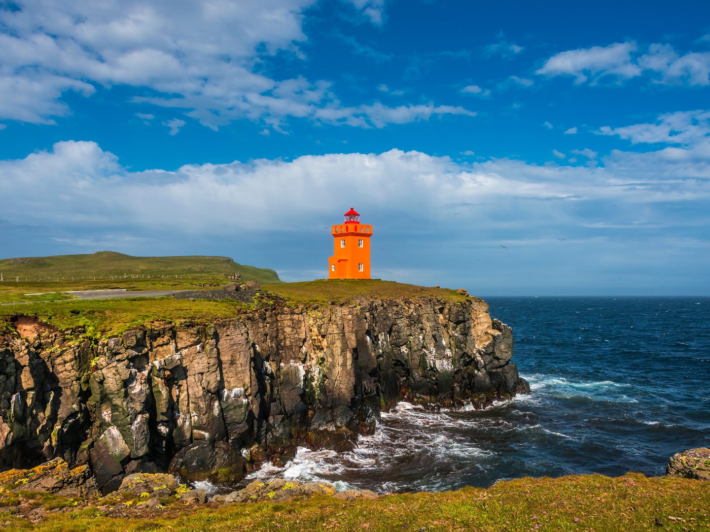 A vibrant orange lighthouse sits atop a rugged cliff against a bright blue sky and turbulent sea. The scene conveys isolation and resilience.