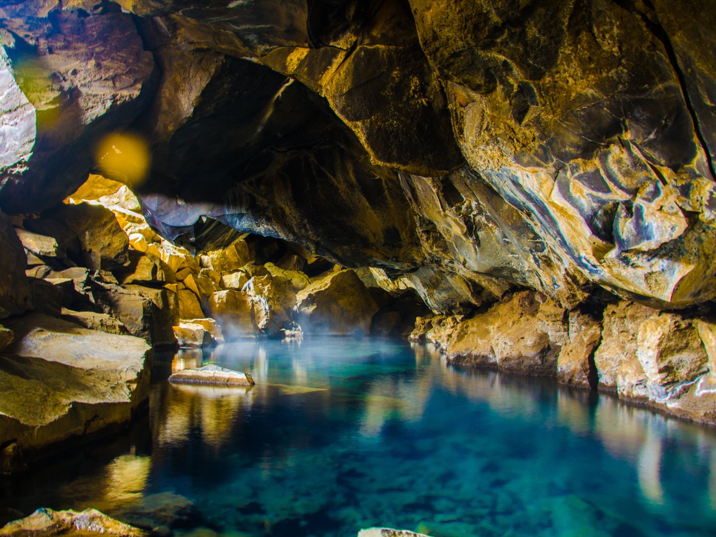A serene cave with turquoise water reflects light off rugged, brown rock formations. Mist hovers above the water, creating a mystical, tranquil atmosphere.
