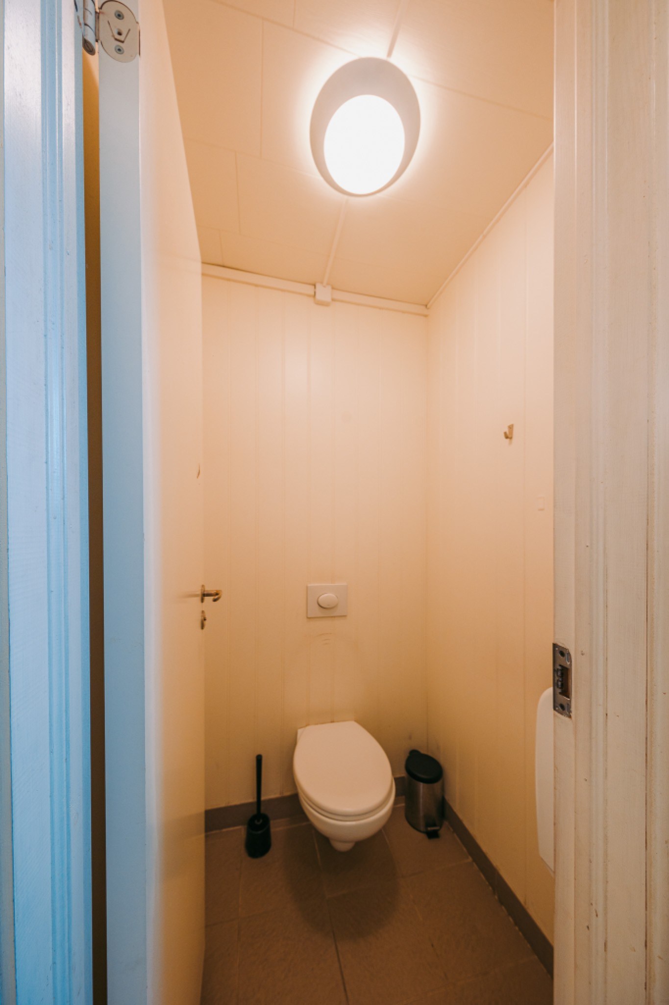 A small, minimal bathroom with a wall-mounted toilet, a black toilet brush, and a silver waste bin.