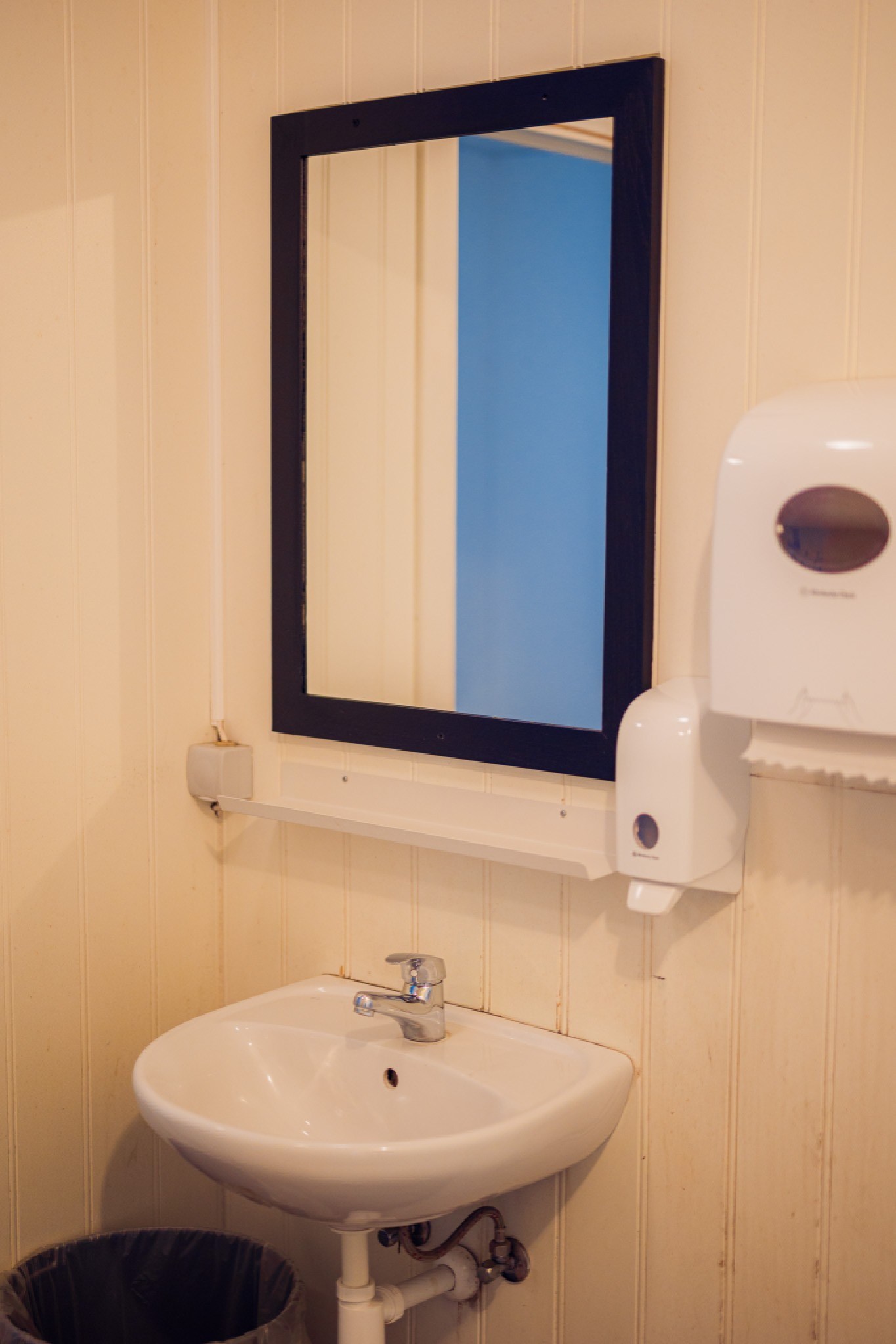 Simple bathroom scene with a white sink beneath a black-framed mirror on cream walls. A white paper towel dispenser is mounted on the right.