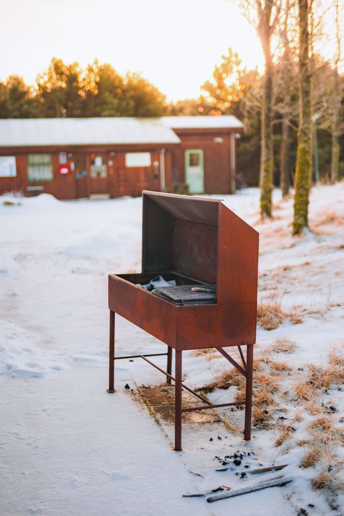 Rustic barbecue grill on snowy ground near a wooden cabin and trees at sunset.