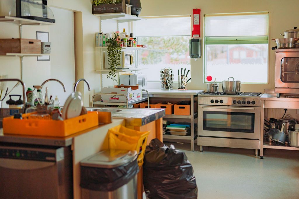 A cozy kitchen with a stainless steel stove and oven, countertop covered in dishes and cleaning supplies, shelves with spices and pots, all under warm lighting.