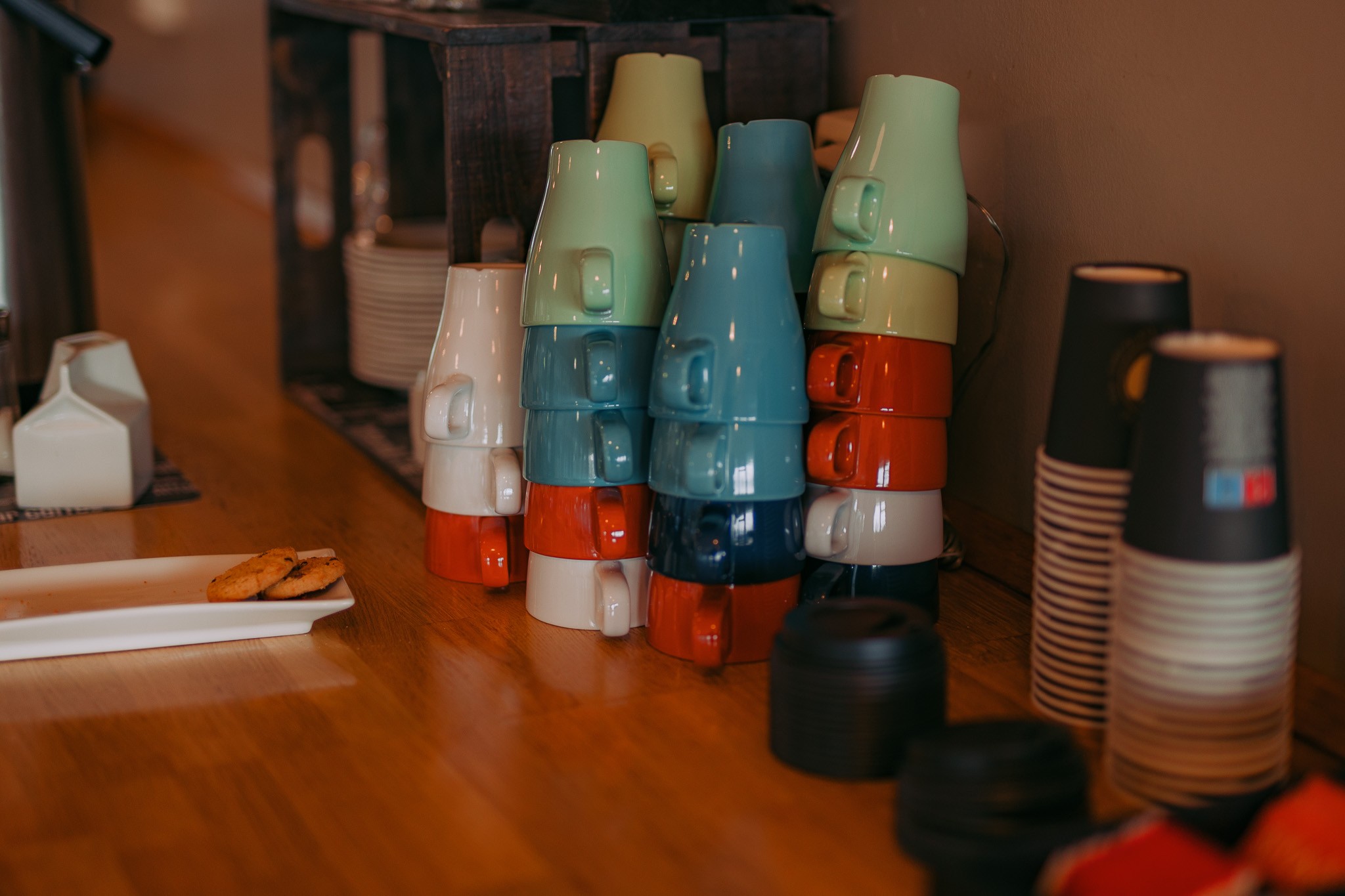 Stacked colorful mugs on a wooden counter beside takeaway cups and two cookies on a plate.