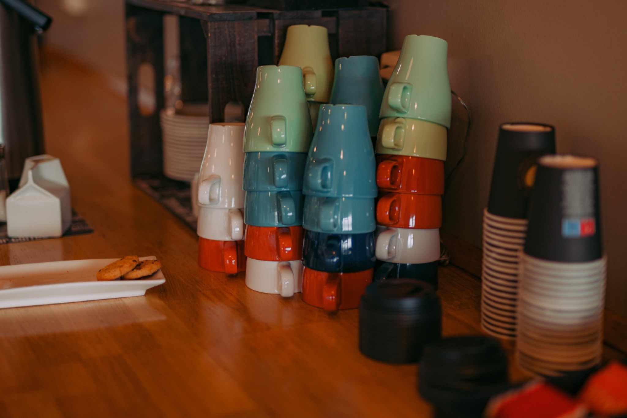 Stacked colorful mugs on a wooden counter beside takeaway cups and two cookies on a plate.