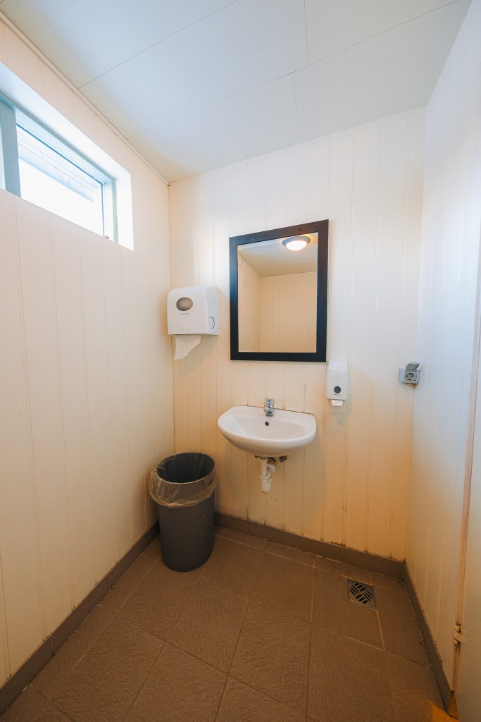 Compact bathroom with white paneled walls, a small sink, a black-framed mirror, and a paper towel dispenser. A trash can sits nearby on a tiled floor.