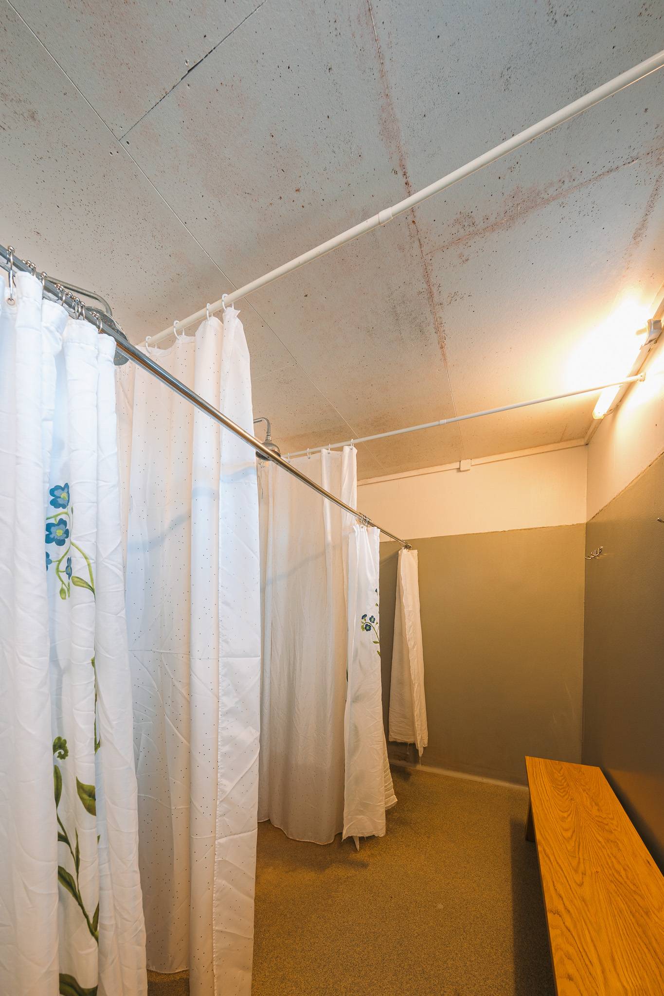 Dimly lit locker room with white shower curtains featuring blue floral designs. Brown bench on the right.