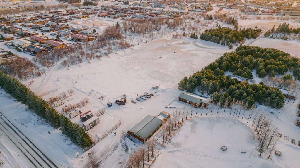 Aerial view of a campground with buildings, trees, and several cars, with the town of Selfoss in the background.